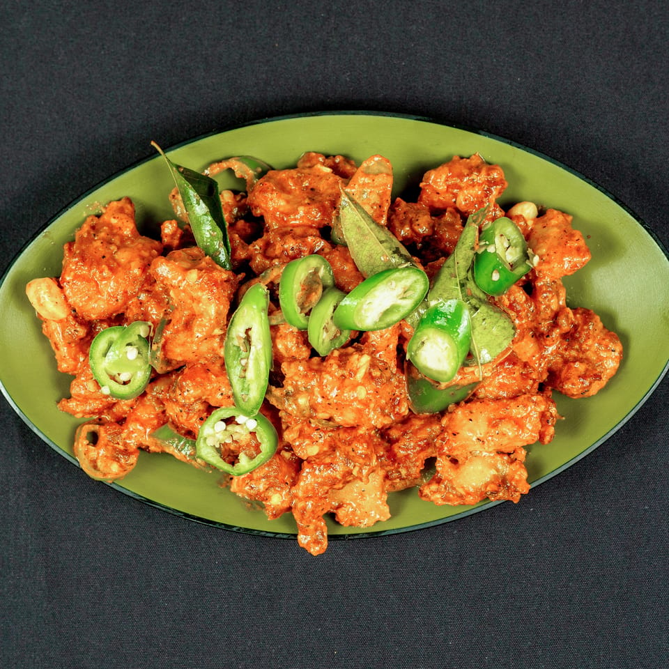Crispy orange-coated fried pieces garnished with sliced green chilies and curry leaves on a green oval plate.
