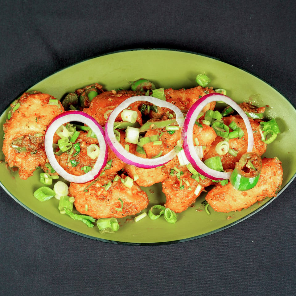 Spiced golden fried bites garnished with red onion rings, green chilies, scallions, and herbs on a green platter.