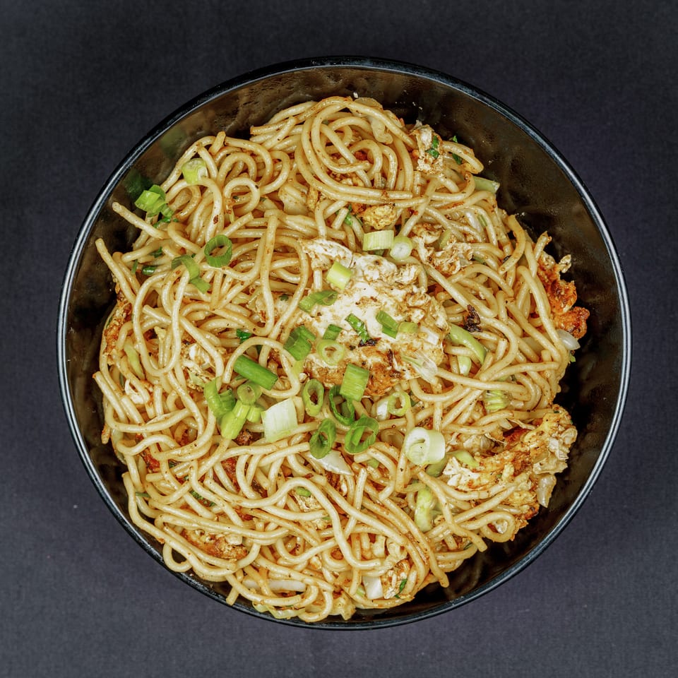 Bowl of seasoned noodles with scrambled egg, vegetables, and sliced green onions on a dark surface.