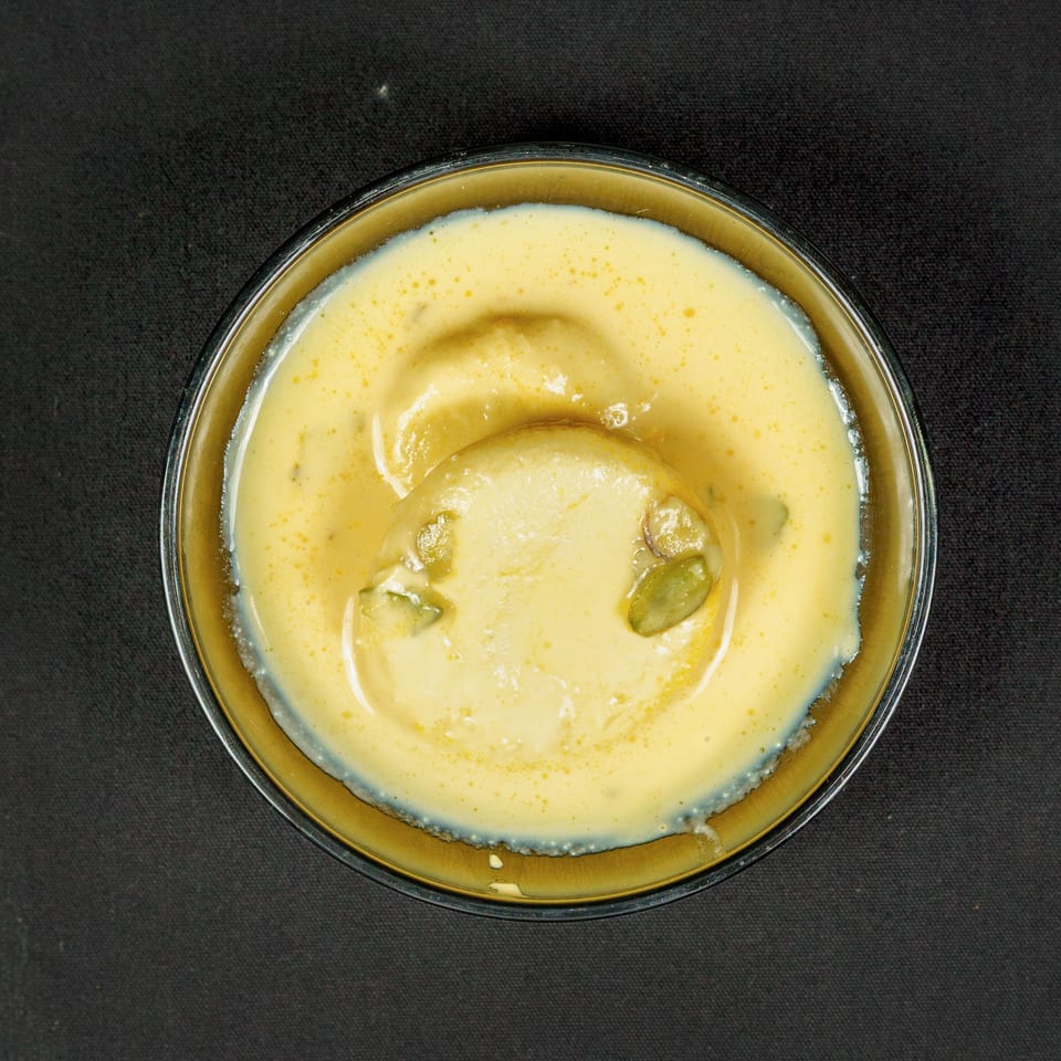 Top-down view of a creamy yellow dessert topped with green pistachios in a glass bowl on black fabric.