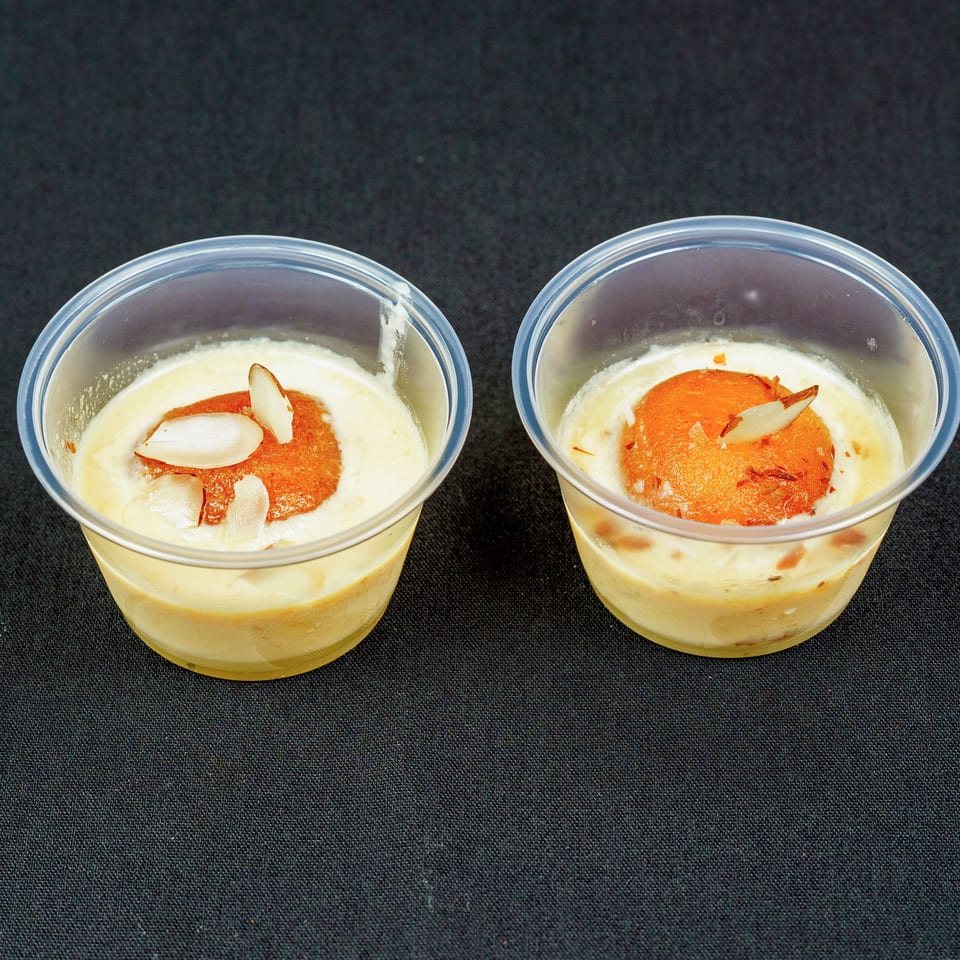 Two clear cups of creamy dessert topped with orange-colored sweets and almond slivers on black fabric.