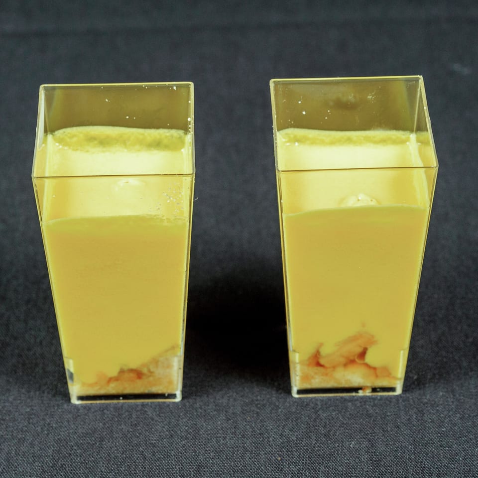 Two square glasses of yellow layered dessert with fruit pieces at the bottom, set against a dark background.