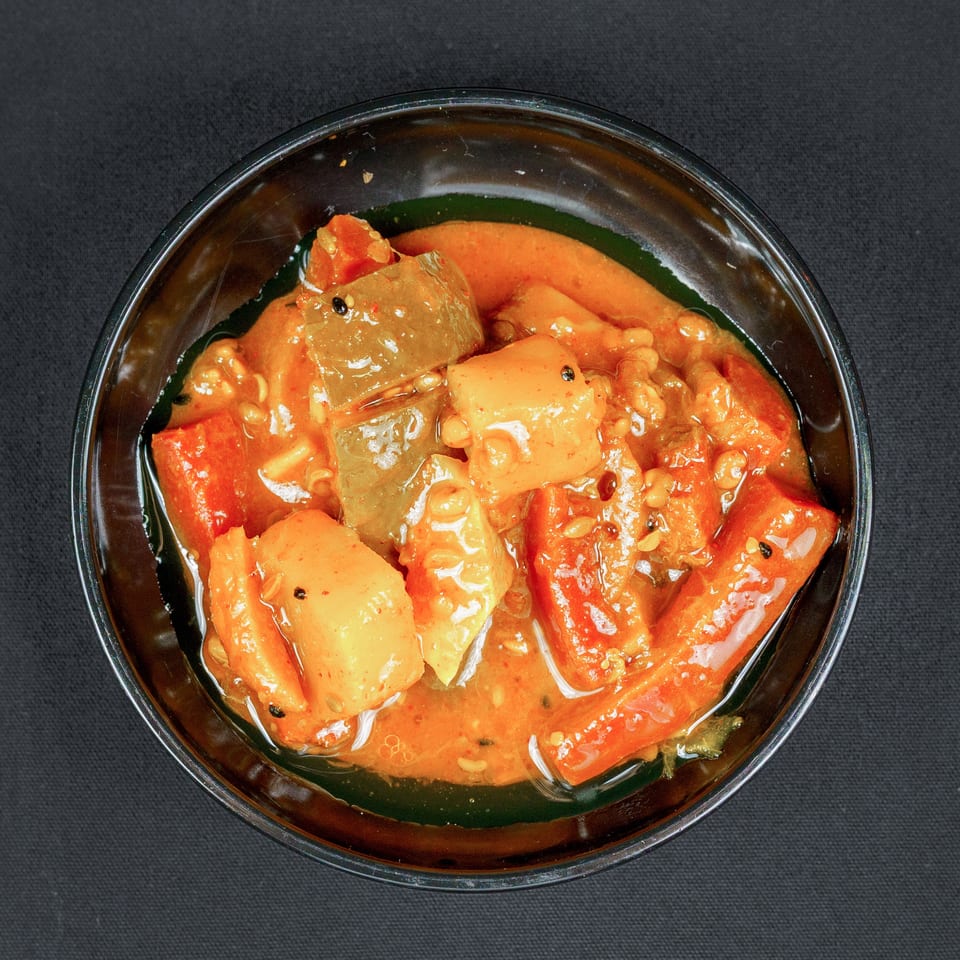 Overhead view of chunky vegetables and spices in a glossy red-orange sauce, served in a glass bowl on black fabric.