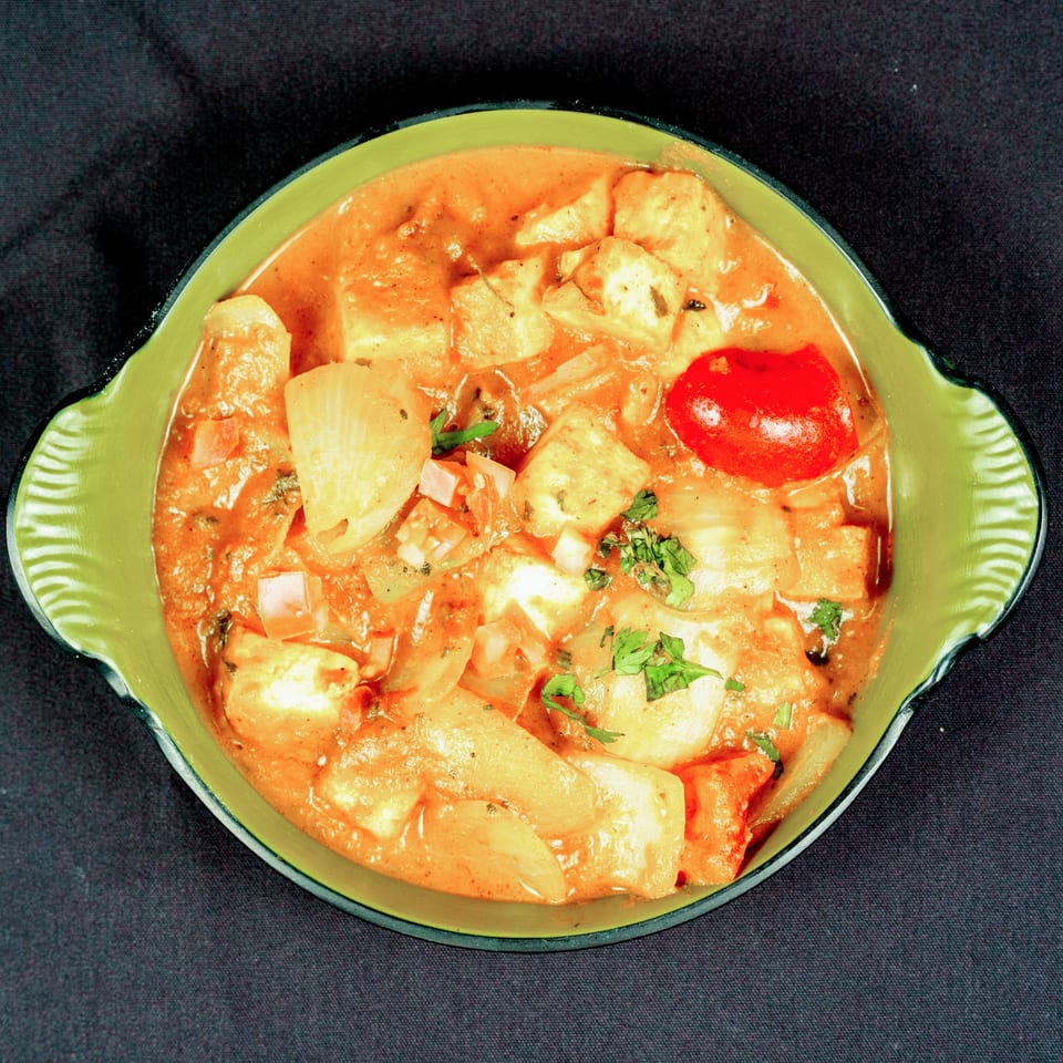 Bowl of creamy orange curry with paneer, vegetables, tomato, and cilantro garnish on a dark surface.